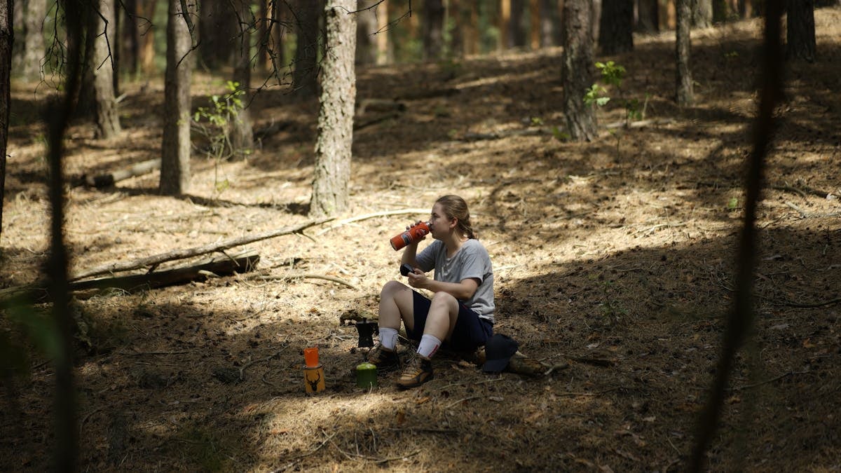 A woman relaxes in a sunny forest, enjoying a drink and solitude.